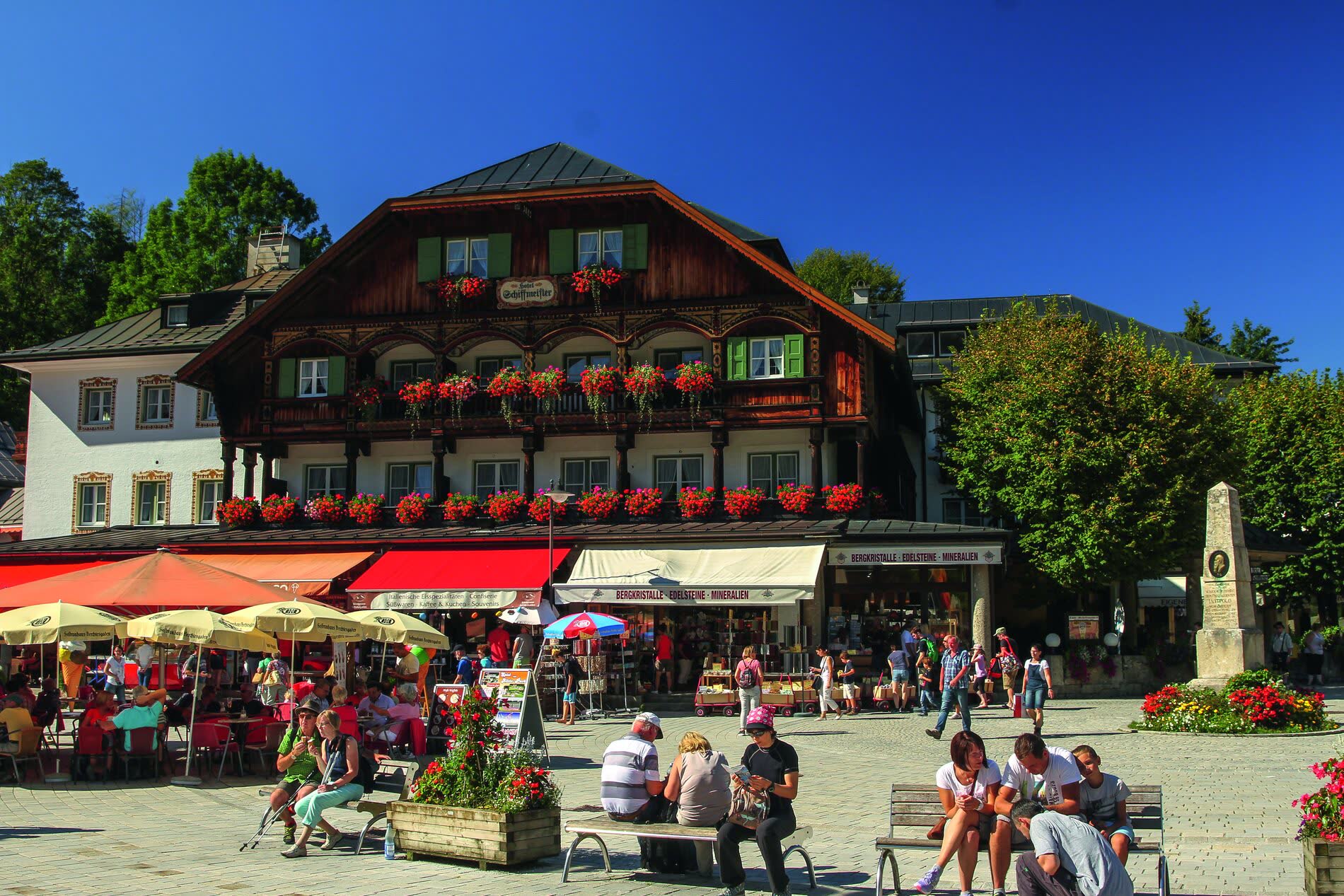 Holzhaus mit geschnitzten Balkonen und roten Geranien, davor Sonnenschirme und Besucher auf einem Platz Holzhaus mit geschnitzten Balkonen und roten Geranien, davor Sonnenschirme und Besucher auf einem Platz